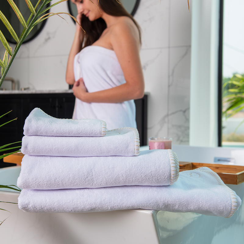 Brunette model wearing a bath towel in a marble bathroom with 4 white nautical themed towels folded in foreground.