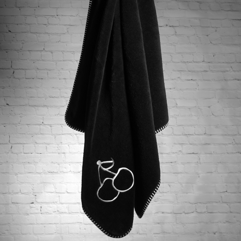 A black nautical themed towel with white stitching and an embroidered image of two cherries, hanging in front of a wall of white bricks.