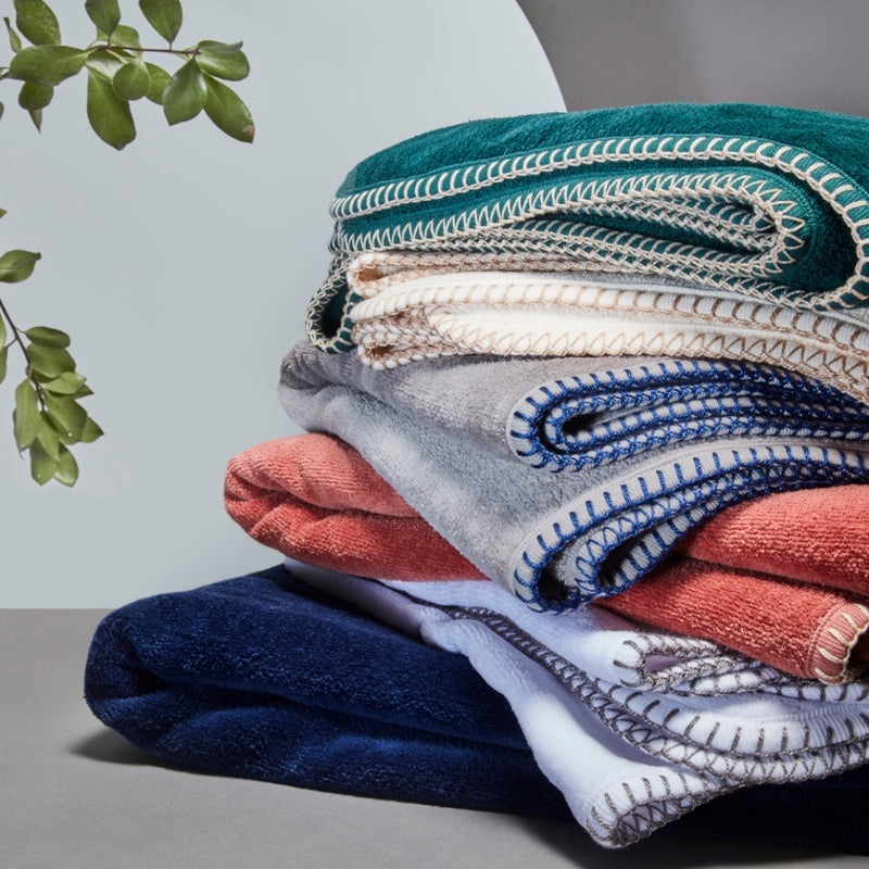 A stck of 6 nautical themed bath towels in blue, white, terracotta, grey, white and green with colored stitching with a grey and white background and leaves of a plant