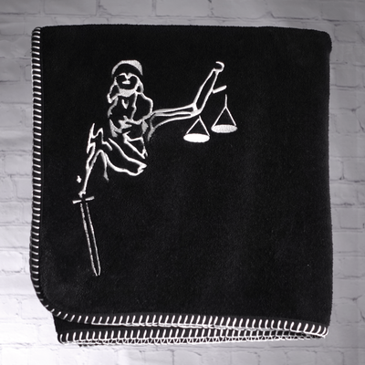 A black nautical themed towel with white edge stitching and an image of Lady Justice holding the scales of justice, embriodered on it